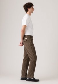 Pantaloni in velluto a coste marrone con taglio dritto, dotati di tasche laterali e toppa posteriore. Indossati con una maglietta bianca semplice e scarpe nere.