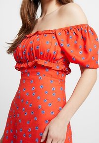 Šaty s odkrytými rameny v jasně oranžové látce. Obsahují malé modré květinové vzory, volánkový výstřih a přiléhavý siluetu.