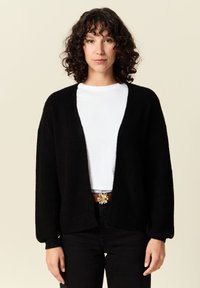 Svart, öppen-front cardigan med strukturerad stickning, slappa ärmar och avslappnad passform, kombinerad med en vit topp och mörka jeans.