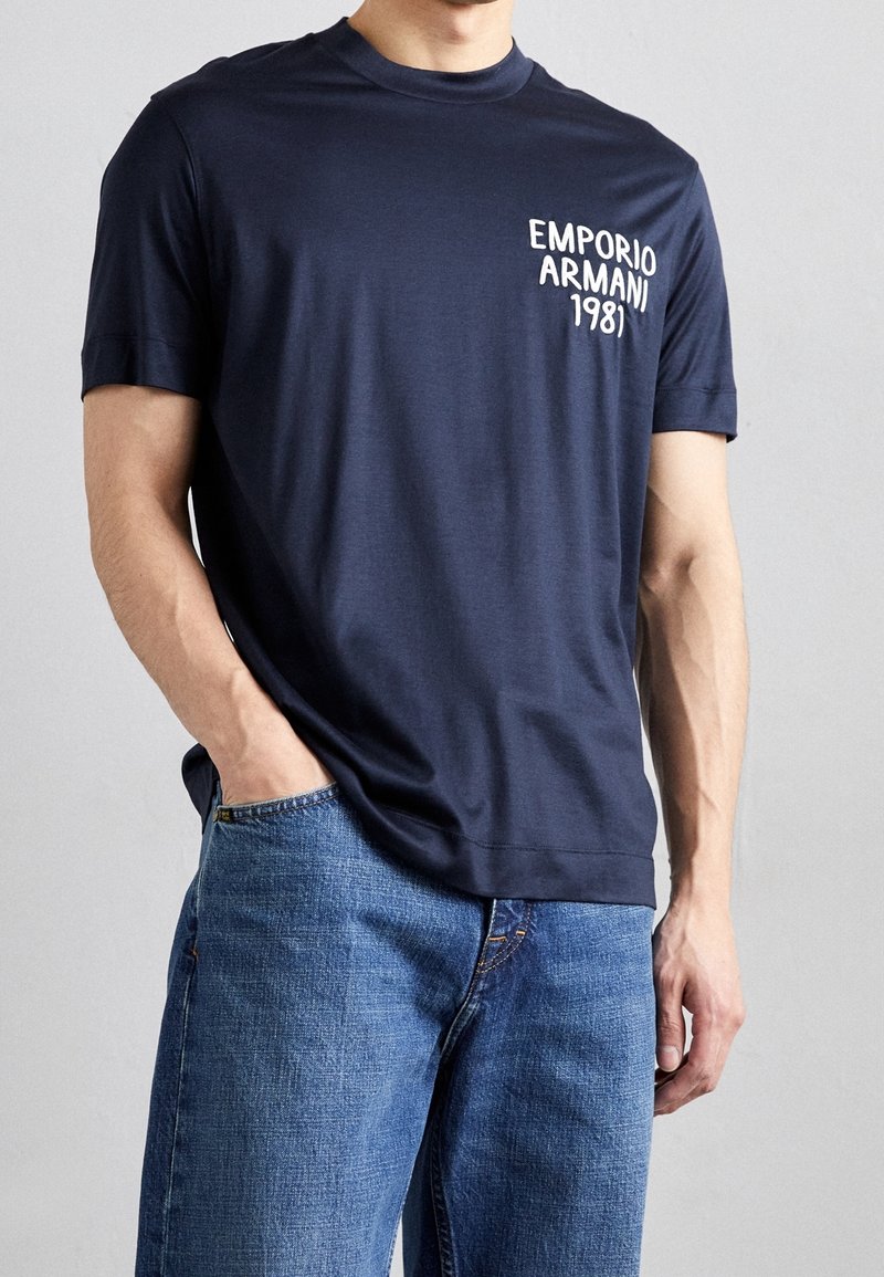 Tmavě modré bavlněné tričko s kulatým výstřihem a krátkými rukávy, s bílým textem "Emporio Armani 1981" na hrudi.