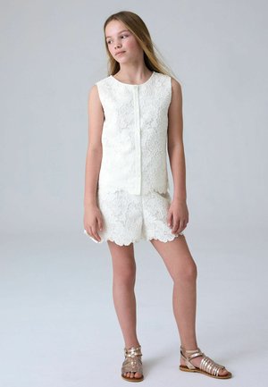 Dievča s dlhými vlasmi nosí biely čipkovaný top bez rukávov a zodpovedajúce šortky, stojí so skríženými nohami a pozerá sa do strany, nosí zlaté sandále.