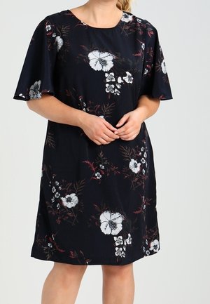 Mujer que lleva un vestido azul marino hasta la rodilla con patrones florales blancos y mangas cortas y anchas, de pie con las manos entrelazadas.