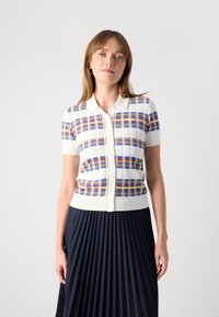 Femme portant un haut à carreaux blanc, bleu et orange à manches courtes, avec un col, associé à une jupe plissée bleu marine.