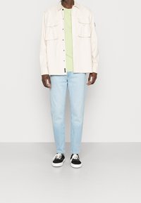 Un homme portant une chemise boutonnée beige clair sur un t-shirt vert clair, un jean bleu clair et des baskets noir et blanc, se tenant sur un sol gris.