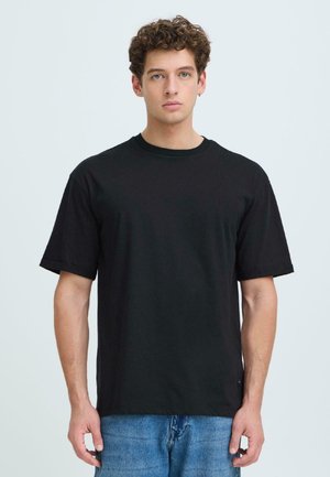 Ung mand med krøllet hår iført en ensfarvet sort T-shirt og blå jeans, står foran en ensartet lys baggrund.