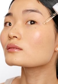 Gesichtsserum-Tropfer wird nahe dem Auge aufgetragen; das Model hat glatte, strahlende Haut, hellrosa Lippen und dunkles Haar. Der Hautton ist hell mit einem subtilen Rouge.
