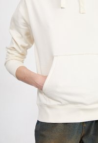 Personne portant un sweat à capuche crème avec une poche avant, main gauche rentrée à l'intérieur, associée à un jean bleu foncé.