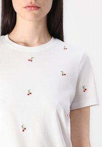 Witte katoenen t-shirt met verspreide rode kersborduursels, korte mouwen en een ronde halslijn. Glad textuur, casual ontwerp.