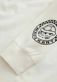 Weißes Langarmshirt mit einem schwarzen Druckdesign, das einen Cartoon-Schildkrötenpanzer zeigt, sowie dem Text "MARIO KART" in einem runden Abzeichen.
