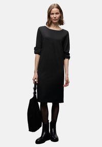 Czarna sukienka z długimi rękawami, okrągłym dekoltem i luźnym krojem. Noszona z czarnymi botkami i teksturowaną czarną torbą typu tote. Gładka tkanina.