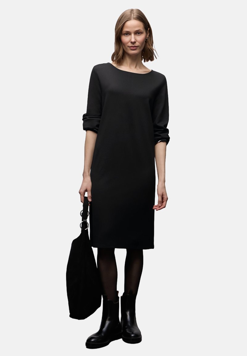 Czarna sukienka z długimi rękawami, okrągłym dekoltem i luźnym krojem. Noszona z czarnymi botkami i teksturowaną czarną torbą typu tote. Gładka tkanina.