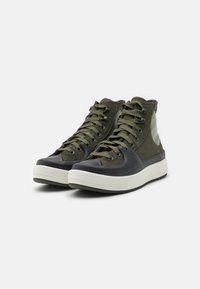 Sneakers typu high-top w oliwkowej tkaninie, gumowy czubek, teksturowana powierzchnia oraz kontrastowa biała gumowa podeszwa. Sznurowane pasującymi zielonymi sznurówkami.