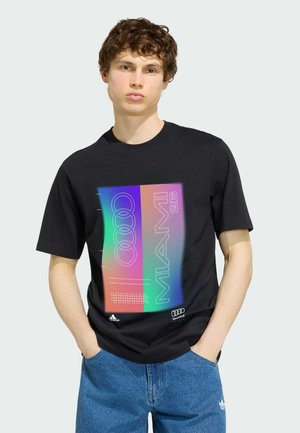 Młody mężczyzna o kręconych włosach, ubrany w czarną koszulkę z kolorową grafiką i logotypem "Miami 26", w połączeniu z niebieskimi dżinsami, ręce w kieszeniach.