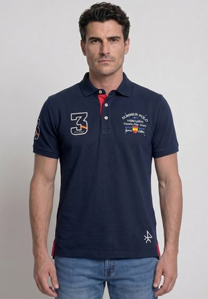 Hombre con camiseta polo azul marino con los logotipos "3" y "Summer Polo 1971 España Polo Team", de pie frente a un fondo liso.