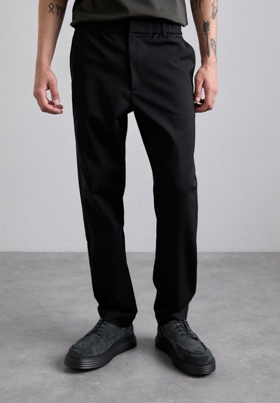 Schwarze, maßgeschneiderte Hosen mit einem elastischen Bund, schmal zulaufenden Beinen und einer glatten Stoffstruktur, kombiniert mit dunklen, schnürbaren Schuhen.