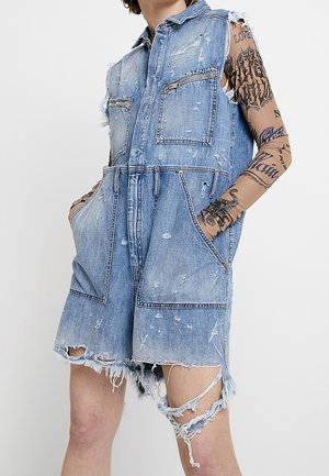 Combinaison sans manches en denim usé avec poches avant, portée sur des bras tatoués, présentant des ourlets effilochés et des détails déchirés sur les jambes.