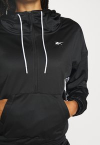 Personne portant un sweat à capuche Reebok noir à demi-fermeture éclair avec des cordons blancs et les mains dans la poche avant, sur un fond uni.