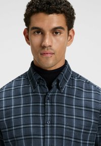 Blauwe geruite button-up shirt met een kraagdesign, met subtiele grijze lijnen en donkere knopen. Draagt over een zwarte turtleneck.
