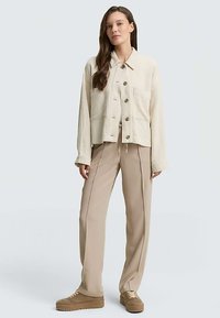 Veste beige boutonnée avec deux poches avant, associée à un pantalon beige et des baskets marron. Lignes épurées, coupe décontractée, tissu léger.