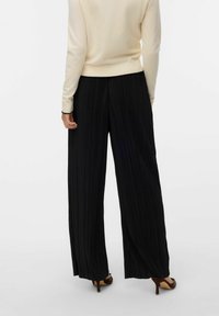 Svarta plisserade vida byxor med hög midja, kombinerade med en ljusbeige tröja. Slät textur med vertikala ränder.