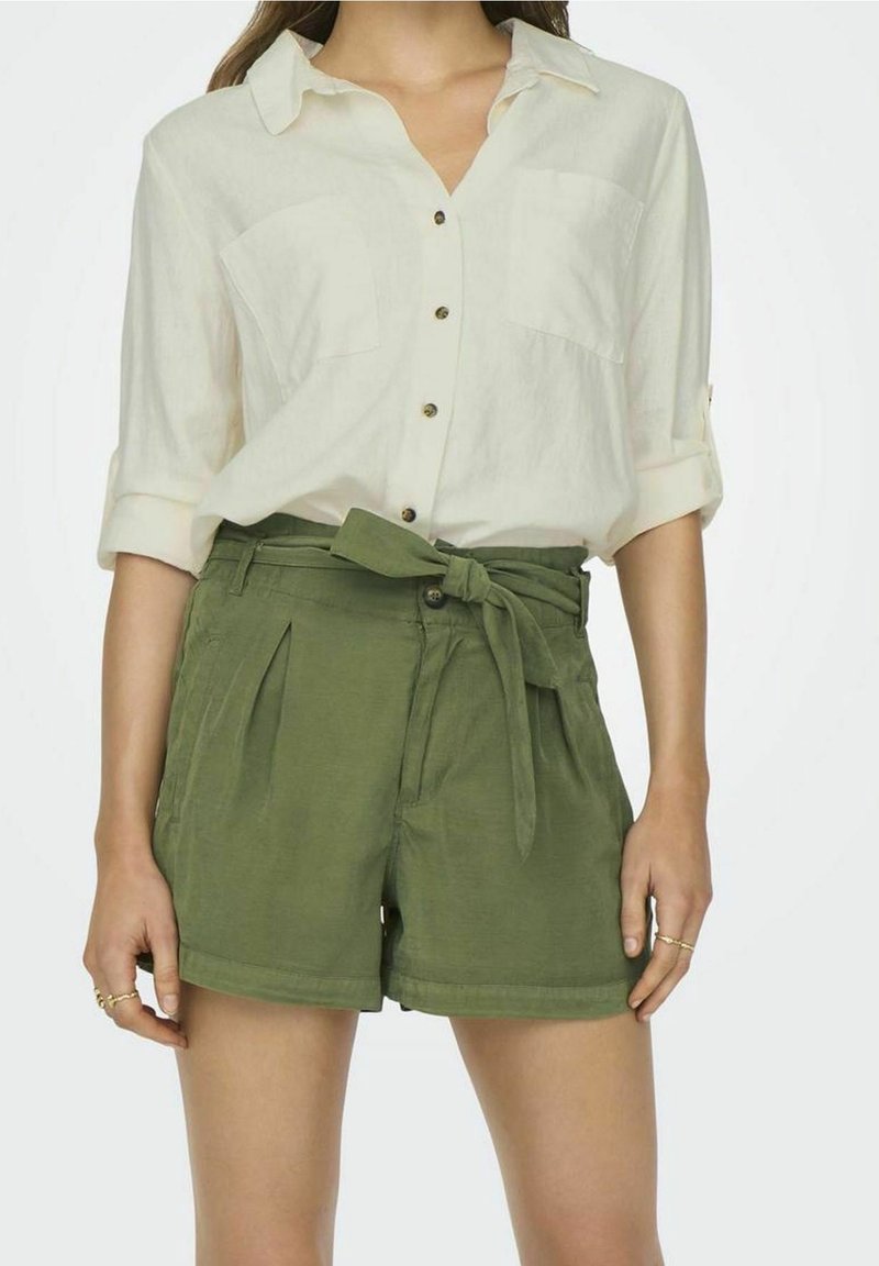 Femme portant une chemise blanche boutonnée avec les manches retroussées et un short vert taille haute noué avec une ceinture en tissu.