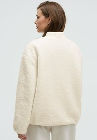 Cream-colored fleece jacket with a high collar and loose fit. Soft, textured surface; dropped shoulders and wide sleeves for comfort.