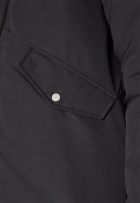 Chaqueta negra con una tela texturizada, que presenta un cierre de botón y un bolsillo diagonal y en ángulo en la parte delantera, con detalles de costura evidentes.