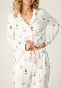 Vit pyjamaset med ett champagneflask- och blommönster. Tröjan har rosa kantband och knappar; byxorna har en rosa dragsko. Mjuk tyg.