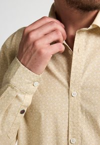 Camisa de un amarillo claro con un sutil diseño geométrico, cuello abotonado y acentos de botones en contraste en los puños.