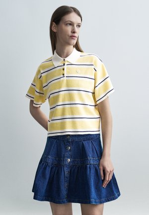 Mladá žena v žlto-bielom prúžkovanom krátkoročnom polokošeli a modrej džínsovej sukni s gombíkmi, stojaca s rukou na bokoch.