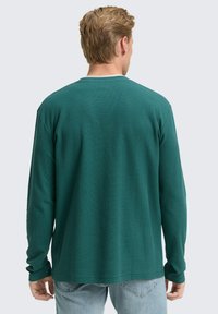 Man met kort lichtbruin haar, gekleed in een donkergroene textuurhemd met lange mouwen en lichtblauwe jeans, met de rug naar de camera op een effen achtergrond.
