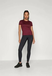 Burgundijas krāsu pieguļošs T-krekls ar īsām piedurknēm, kombinēts ar augsto vidukli melnām biksēm un melniem sporta apaviem, neitrālā fonā.