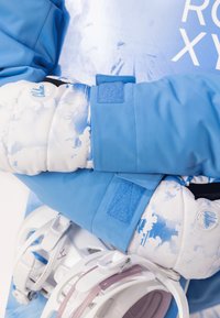 Blå skidjacka med vita och ljusblå molnmönster, Velcro-detallar, i kombination med en vit snowboardbindning.