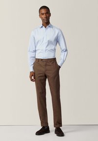 Camisa azul claro de botones, pantalones marrones con textura, zapatos oscuros. La camisa tiene un cuello estándar y mangas largas, metida de manera ordenada por dentro.