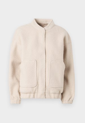 Beige fliistekampsun kõrge kaelusega, kahe eesmine taskuga ja elastsete varrukatega. Pehme tekstuur, kastja siluett ja minimaalne detaile.