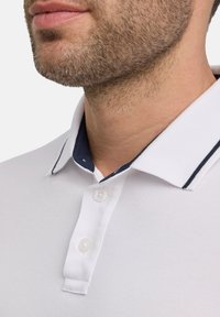 Biała koszulka polo z granatowym kołnierzykiem z detalami w paski i trzema guzikami, wykonana z teksturowanej tkaniny. Zbliżenie na kołnierz i obszar szyi.