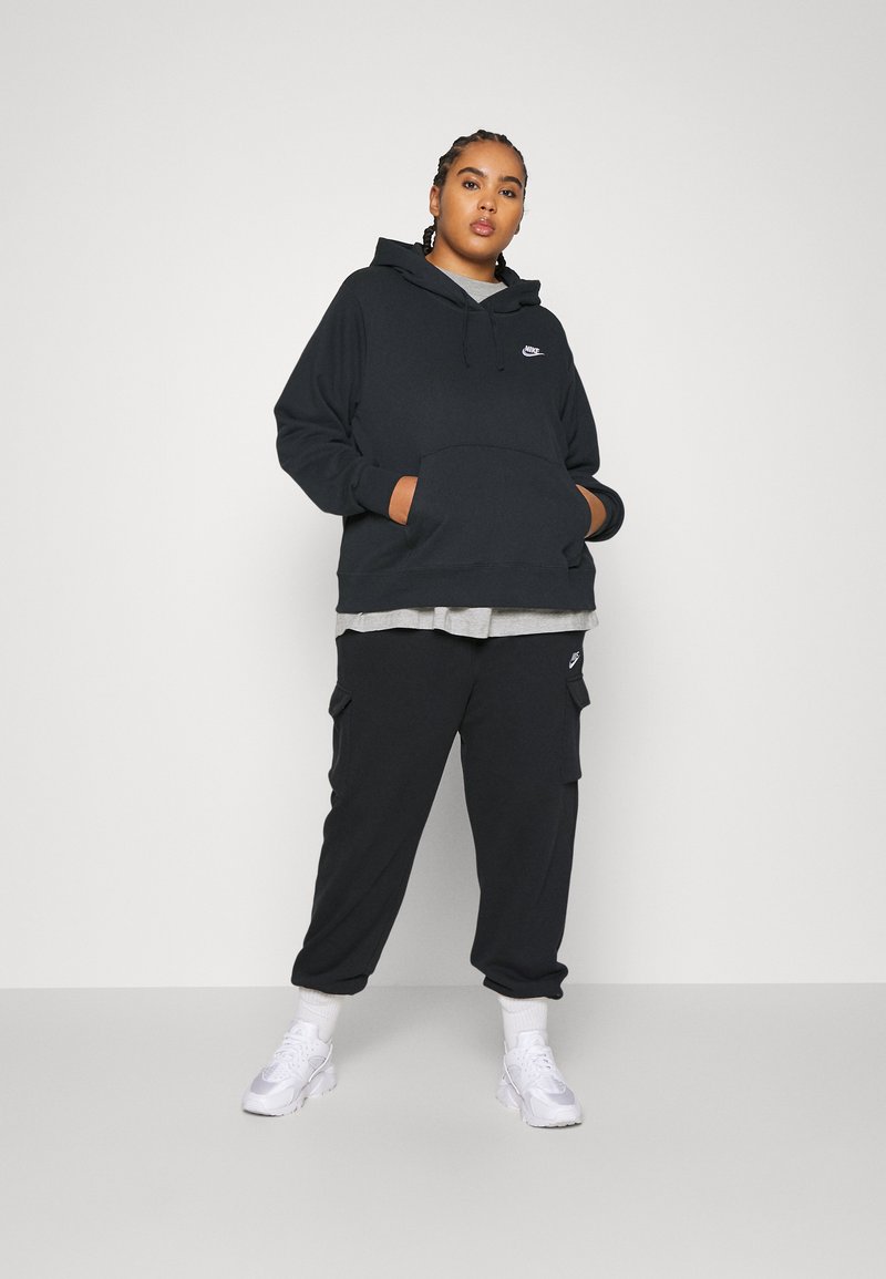 Sudadera negra de gran tamaño con un bolsillo frontal y un logo bordado, combinada con pantalones cargo negros. Zapatillas blancas complementan el conjunto.