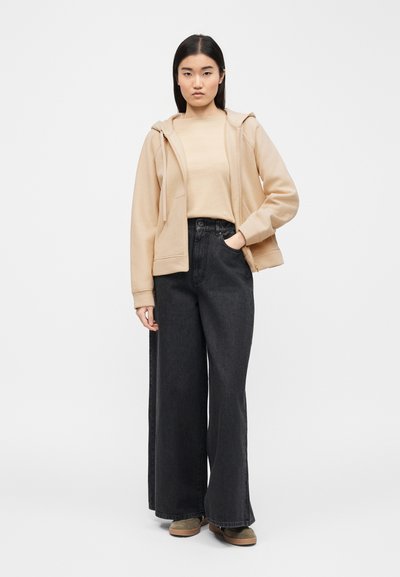 Γυναίκα που στέκεται, φοράει μπεζ φούτερ με φερμουάρ, αντίστοιχο μπεζ πουκάμισο, φαρδιά μαύρα τζιν παντελόνια και καφέ παπούτσια, σε απλό λευκό φόντο.