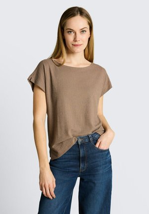 Jonge vrouw met lang haar draagt een losjes vallend bruin gestructureerd kort mouwtopje en donkerblauwe jeans met hoge taille, poseert met één hand in de zak.