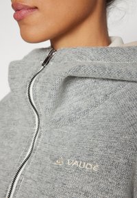 Gray zip-up jacket with a textured knit fabric, featuring a high collar and contrasting zipper, with a subtle logo on the chest.