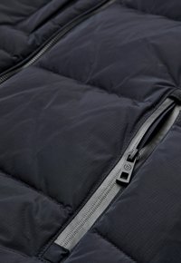 Schwarze Daunenjacke mit großen gesteppten Paneelen, glatter Textur und einem grauen Reißverschluss mit Zugschlaufe zum Schließen.