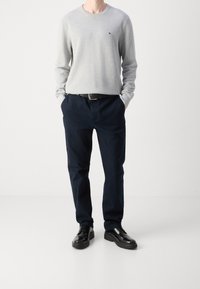 Grauer langärmeliger Pullover, marineblaue Hose, schwarzer Gürtel und schwarze Schuhe. Der Pullover hat einen Rundhalsausschnitt; die Hose ist gerade geschnitten.