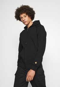 Schwarze Zip-Hoodie aus Baumwollgemisch, mit einer Vorderseite-Tasche und goldenen Logo-Akzenten am Ärmel. Lässige Passform mit Kapuze.