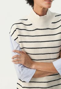 Frau, die einen cremefarbenen und schwarzen gestreiften gestrickten ärmellosen Oberteil über einem hellblauen Langarmshirt trägt und die Arme verschränkt hat.