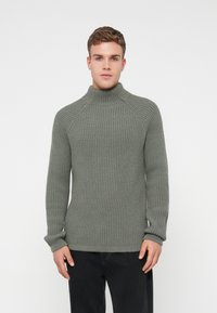 Pelēka ribota adīta augsta apkakle ar raglanu piedurknēm un teksturētu apdari. Apvienota ar tumšām džinsām, izceļot slaidu piegriezumu.