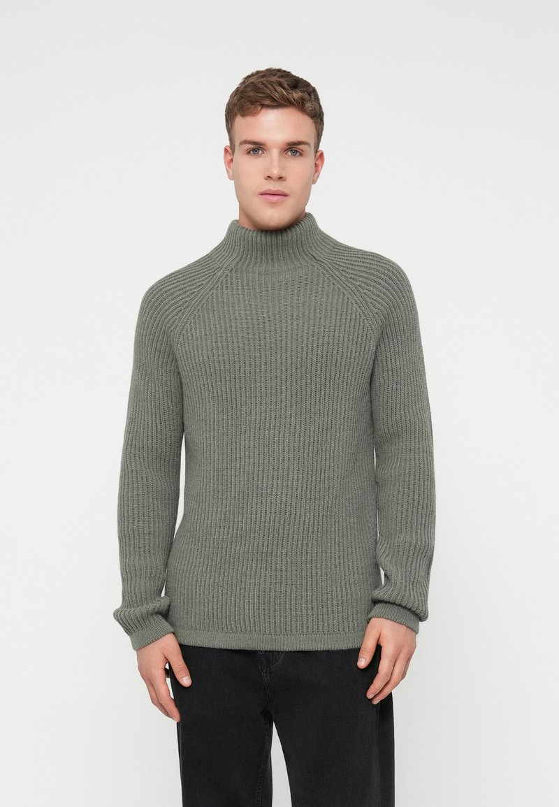 Pelēka ribota adīta augsta apkakle ar raglanu piedurknēm un teksturētu apdari. Apvienota ar tumšām džinsām, izceļot slaidu piegriezumu.