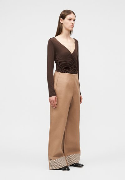 Γυναίκα που φοράει καφέ ρούχα με μακρυά μανίκια και ρέλια, με βαθύ V στο ντεκολτέ, μπεζ παντελόνι άνετης γραμμής με καρό μανσέτες και μαύρα μυτερά παπούτσια.