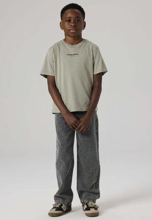 Young boy standing with hands clasped, wearing a light green t-shirt, gray jeans, and black-and-beige sneakers on a plain background.