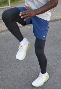 Leggings atléticos negros, pantalones cortos azules, zapatillas blancas con suelas verdes y una camiseta gris. La persona está en una pose dinámica de ejercicio.