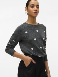Szürke kötött pulóver fehér szívmintákkal, kerek nyakkivágással és hosszú ujjal. A modell fekete nadrágot visel, kezét a zsebében tartja.